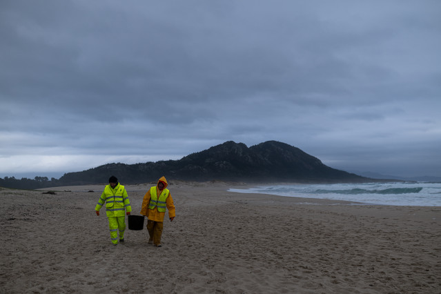 Dos hombres cargan un cubo lleno de pélets, en la playa Area Maior, a 13 de enero de 2024, en Muros, A Coruña, Galicia.