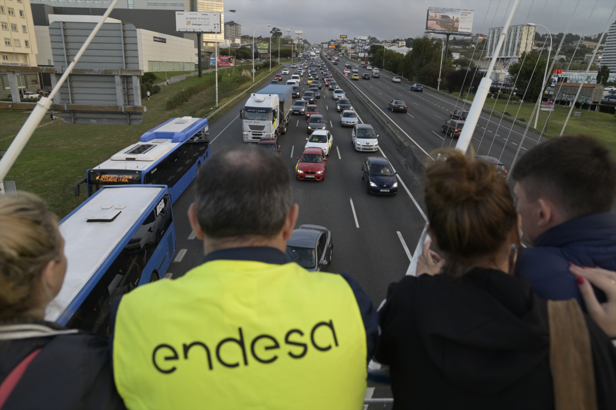 Varias personas, una con un chaleco de 'Endesa', observan una caravana de automóviles y camiones movilizada por la paralización de la central térmica de Endesa en de As Pontes se dirige desde esa l