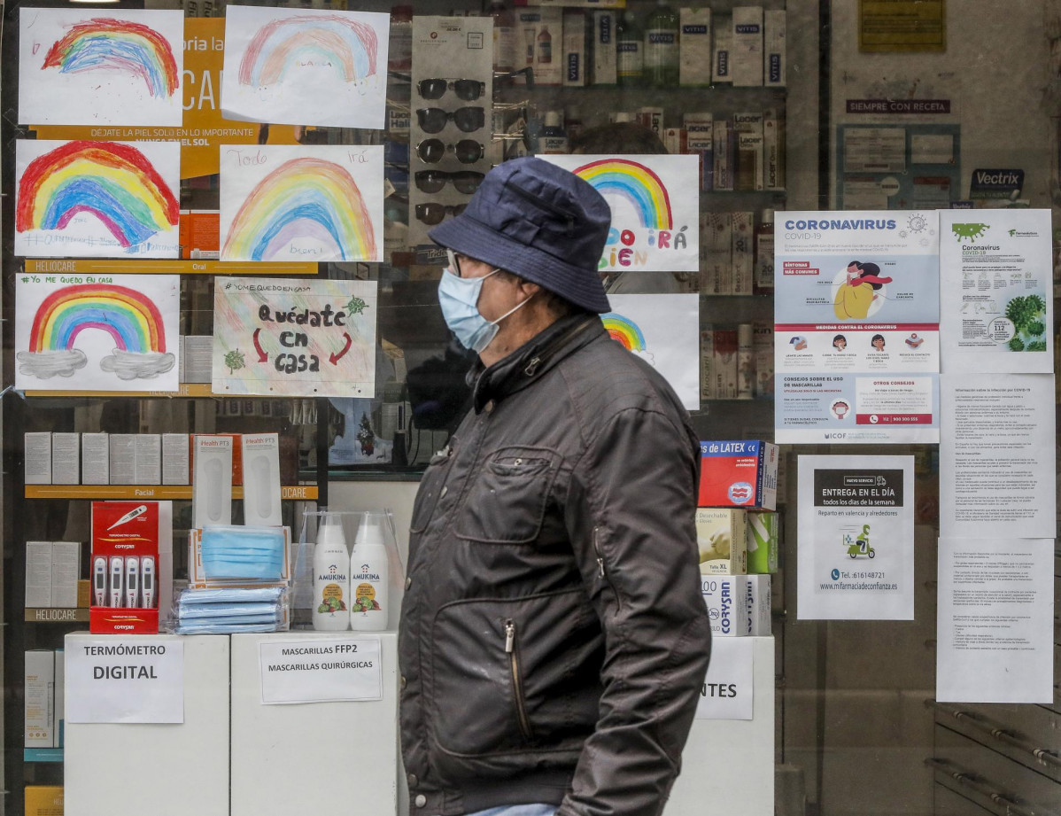 Un hombre con mascarilla pasa junto a una farmacia con dibujos infantiles en su escaparate .