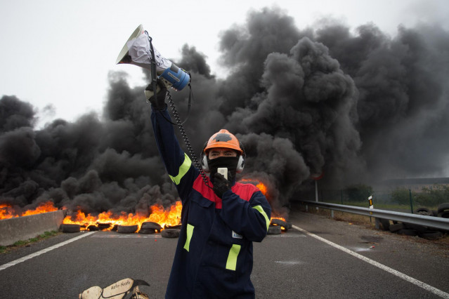 Un participante de la manifestación del comité de empresa de Alcoa habla por un megáfono junto a la quema de unos neumáticos tras el anuncio hace tres días del despido colectivo a 31 de mayo de 2020