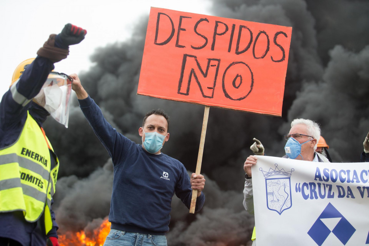Un participante de la manifestación del comité de empresa de Alcoa lleva un cartel en el que se lee 'Despidos no' junto a la quema de unos neumáticos tras el anuncio hace tres días del despido. En